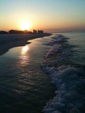 A beach scene with the sun setting over the ocean, waves gently lapping at the shore, and a pier extending into the water. - Last minute discounts - Steps to the Bay - Baywatch A1 (Pensacola Beach)
