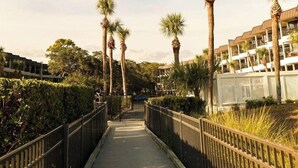 A wooden boardwalk with palm trees, surrounded by greenery and a building with multiple balconies. - On the beach 183 SeaSide Villas (Hilton Head Island)