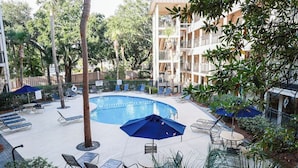A pool area with lounge chairs and umbrellas, surrounded by trees and a multi-story building. - On the beach 183 SeaSide Villas (Hilton Head Island)