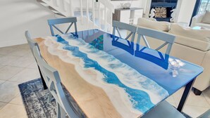 Een eettafel met een strandthema, omringd door stoelen in een moderne woonkamer met een trap en een open haard.