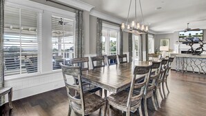 Un espace repas avec une grande table en bois et des chaises assorties, un lustre et une vue sur l’extérieur grâce à de grandes fenêtres.