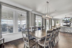 Un comedor con una gran mesa de madera y sillas a juego, una araña y vistas al exterior a través de amplios ventanales.
