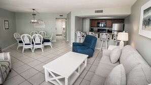Amplia sala de estar con un sofá blanco, un sillón azul y sillas blancas. Se ve una cocina al fondo con electrodomésticos modernos.
