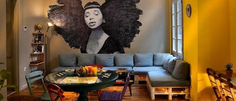 A cozy room with a large mural, a sofa, a dining table with chairs, and a bookshelf.