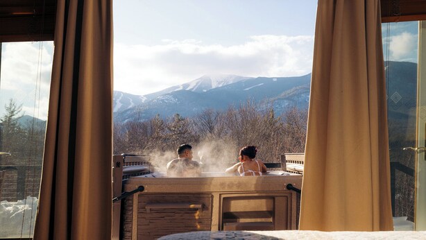 A hot tub with two people enjoying the view of snow-capped mountains through a window with curtains.
