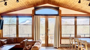 A cozy living room with a high wooden ceiling, large windows offering mountain views, a sofa, a coffee table, and a dining area with chairs.