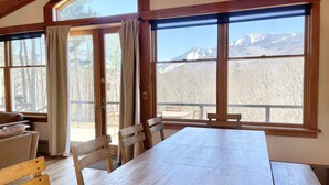 A wooden interior with a large table and chairs, a sofa, and large windows offering mountain views.
