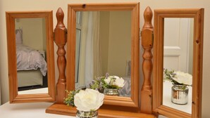 Drei spiegelnde Wände aus Holz, wobei der zentrale Spiegel ein Zimmer mit einem Bett und einer Blumenvase widerspiegelt.