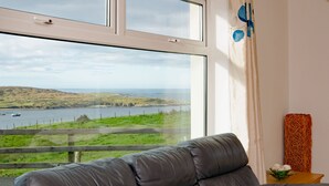 Ein Zimmer mit großem Fenster und Blick auf eine Küstenlandschaft, ein Ledersofa und ein kleiner Tisch mit einer Vase.