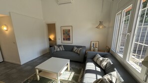 Una sala de estar moderna con un sofá seccional gris, una mesa de centro blanca y un tapete estampado. Hay un cuadro en la pared y una ventana con vistas al exterior.