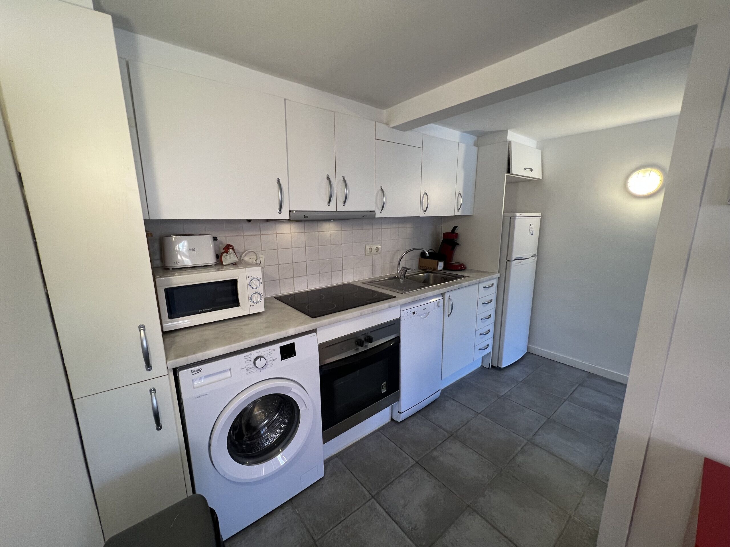 A compact kitchen with white cabinets, a microwave, a toaster, a sink, a dishwasher, and a washing machine.