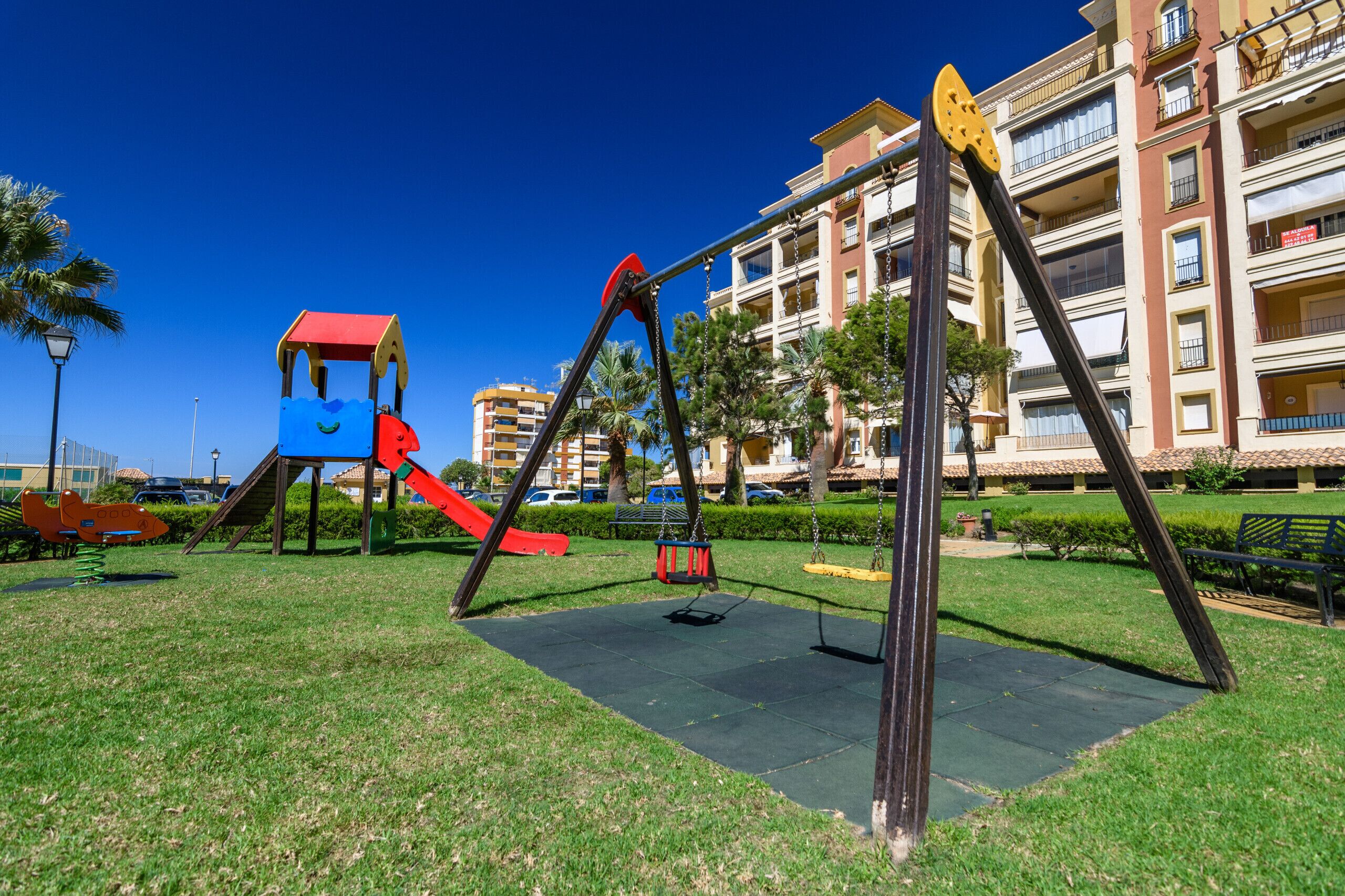 A playground with swings and a slide, surrounded by a grassy area and palm trees, with a building in the background.