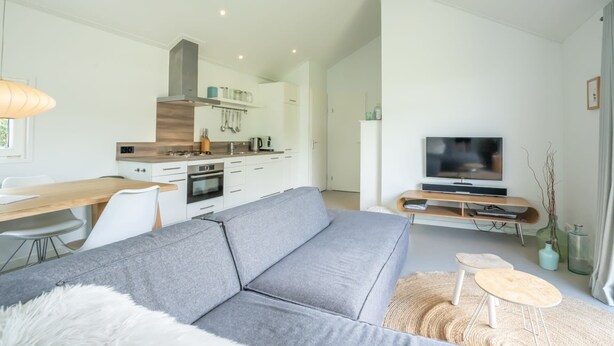 A modern living room with a grey sofa, a dining table, a TV, and a kitchen area in the background.
