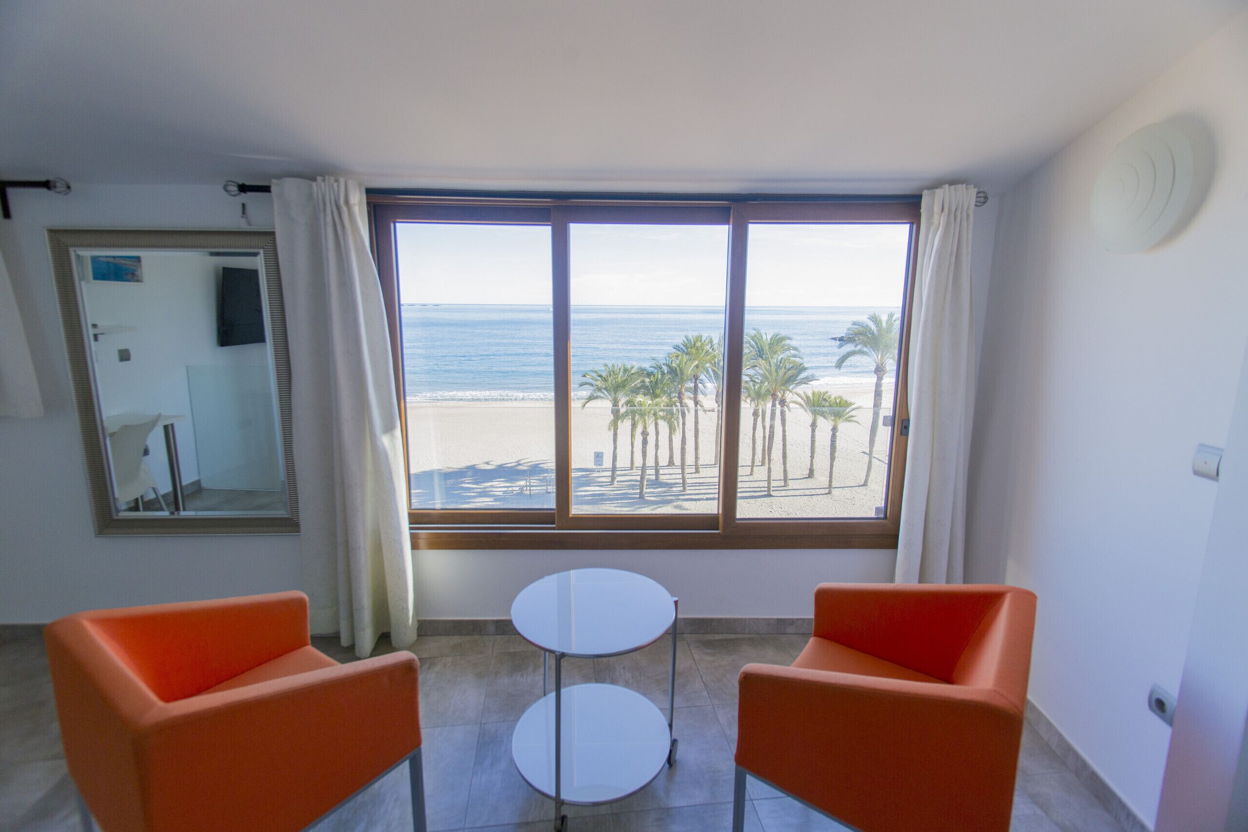 A room with two orange chairs, a small white table, and a large window with a view of the beach and palm trees.