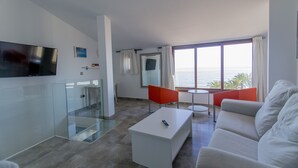 A modern living room with a white sofa, a coffee table, and a TV mounted on the wall. There is a glass partition, a small round table, and a red chair. A large window offers a view of the ocean. - SUPER TORRE - 4 NIVELES CON VISTAS AL MAR (Villajoyosa)