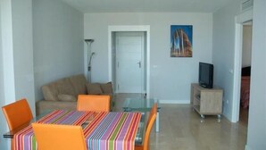 Un espacio habitable compacto con comedor que incluye una mesa de vidrio y sillas naranjas, un sofá, un televisor sobre un mueble bajo y una puerta que da a otra habitación.