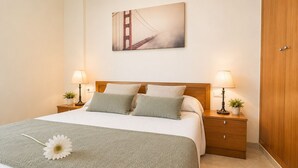 A neatly made bed with a green blanket and pillows, two bedside lamps, a nightstand with a plant, and a painting of a bridge on the wall.