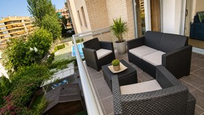 A balcony with wicker furniture, a potted plant, and a view of neighboring buildings and greenery.
