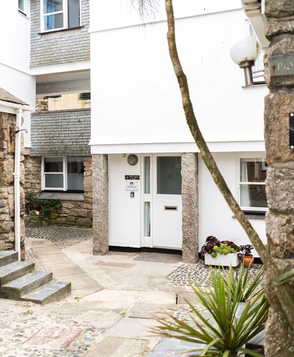A white building with a sign reading "Portsmouth Court." There are stone steps leading to the entrance, a small garden area with plants, and a stone wall on the left.