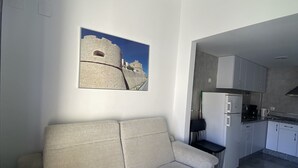 Una sala de estar con un sofá color beige, una cocina con armarios blancos y un cuadro de un edificio histórico en la pared.