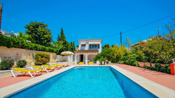 Ein Schwimmbecken mit Liegestühlen, ein Gebäude mit Balkon und umgebende Grünflächen.