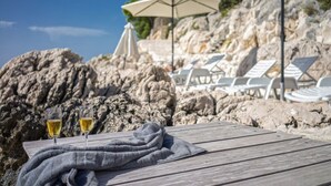 Et træbord med to glas hvidvin, et gråt tæppe og hvide loungesæder samt parasoller i baggrunden.