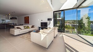 En moderne stue med sofa, sofabord og fjernsyn, et spiseområde med stole, og et køkken med komfur.