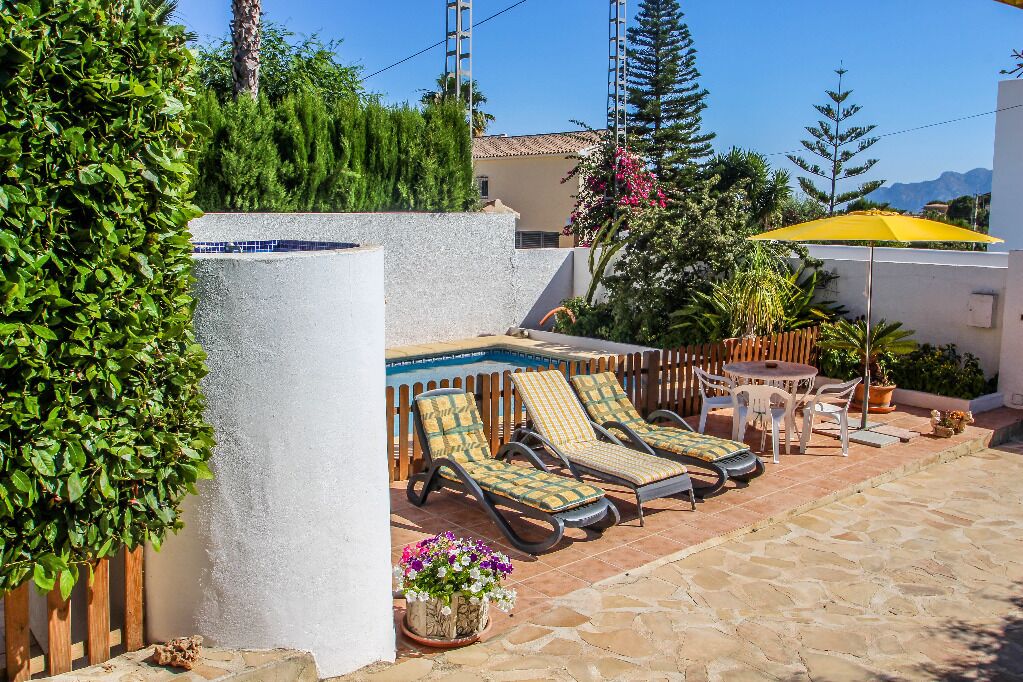 A poolside area with lounge chairs and a small table, surrounded by greenery and a wooden fence.
