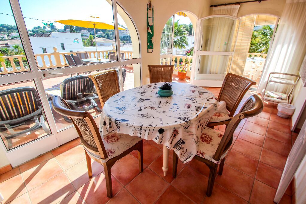 A sunlit patio with a round table and four wicker chairs, a potted plant, and a view of a residential area through large windows.