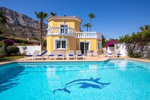 A yellow building with a pool, lounge chairs, and palm trees. - Villa Coral 4 - Denia, Costa Blanca (Denia)