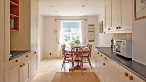 A kitchen with white cabinets, a dining table with chairs, a microwave, and a window with curtains. - Swallows Rest Cottage - sleeps 6 guests in 4 bedrooms (Broadway)