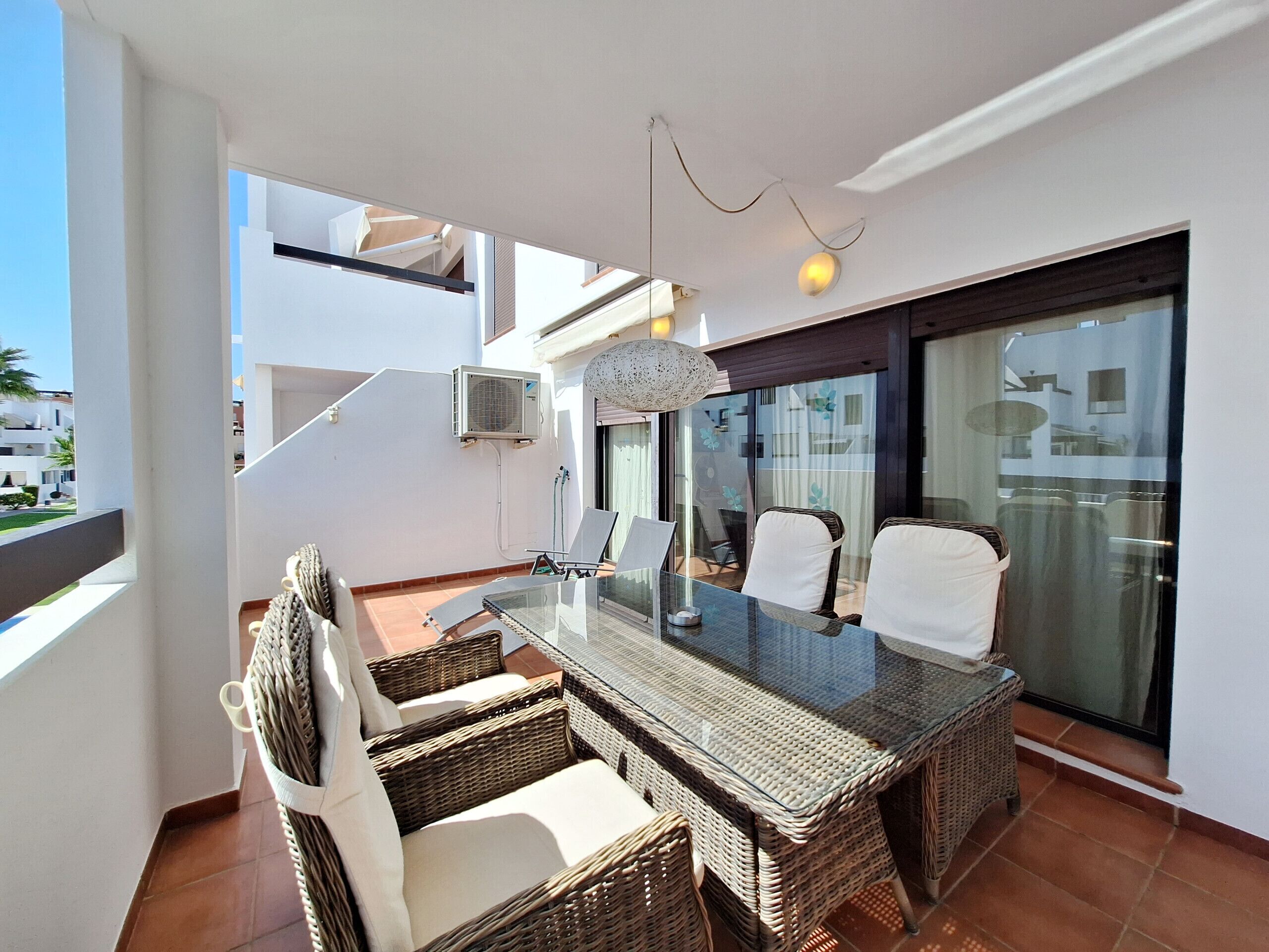 A balcony with wicker furniture, a glass table, and a hanging light fixture.