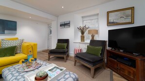 Een woonkamer met een gele bank, twee fauteuils, een salontafel, een televisie en een schilderij aan de muur.