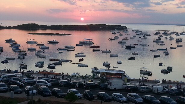 A marina with numerous boats docked, a parking area with cars, and a sunset in the background. - B&B L'Isola (Porto Cesareo)