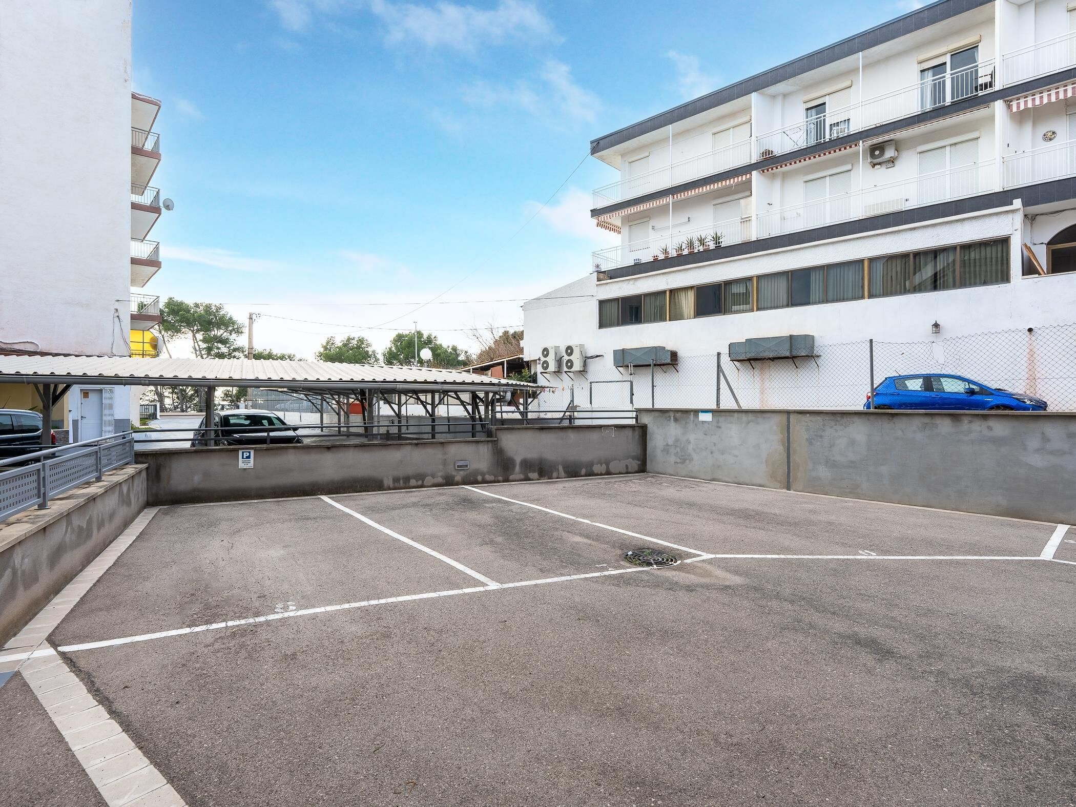 Ein Parkplatz mit überdachtem Bereich, ein blauer Wagen und ein mehrstöckiges Gebäude mit Balkonen.