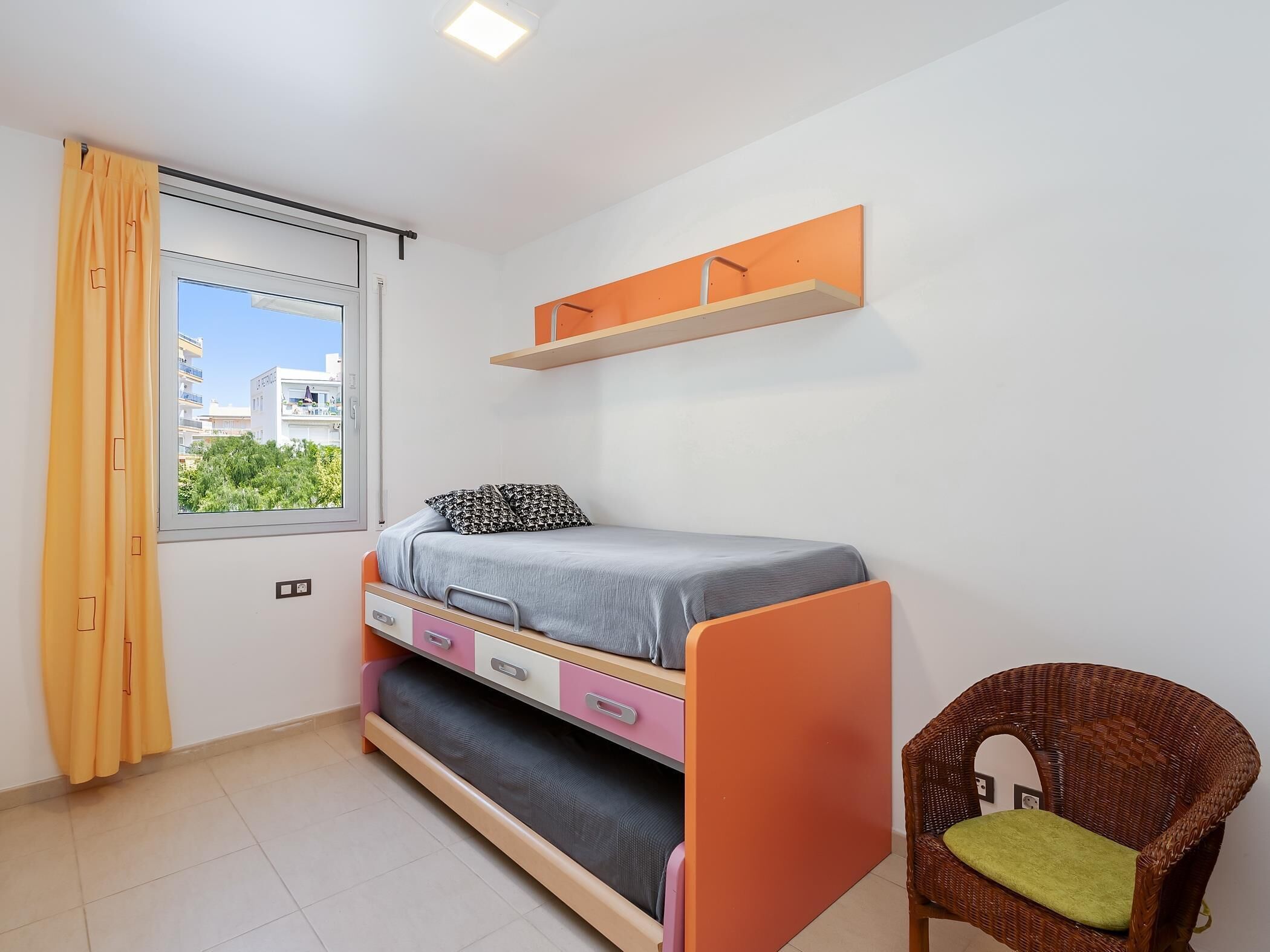 Una habitación pequeña y ordenada con una cama, una silla, una ventana con cortinas y una estantería sobre la cama.