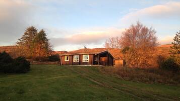A two-story wooden cabin with a large porch, surrounded by a grassy field and trees, set against a backdrop of hills.