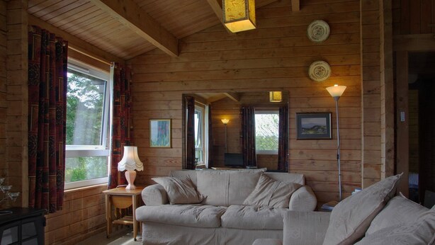 A cozy cabin interior with a wooden interior, a sofa, a lamp, and a window with curtains. - Steading Holidays - Strathconon (Acharacle)