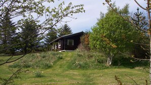 A rustic cabin with a sloped roof, surrounded by a grassy field and dense trees. - Steading Holidays - Strathconon (Acharacle)