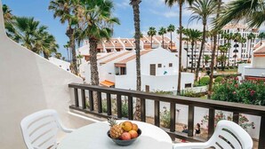 Ein Balkon mit weißen Stühlen und einem Tisch, einer Obstschale und Blick auf Palmen und Gebäude.