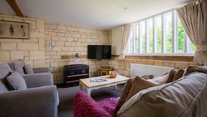 A cozy living room with a stone wall, a television, a wooden coffee table with magazines, and a window with curtains. - A sudeley castle cottage that sleeps 4 guests  in 2 bedrooms (Cheltenham)