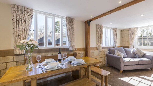 A dining area with a wooden table set for four, a sofa, and a window with curtains. - A sudeley castle cottage that sleeps 4 guests  in 3 bedrooms (Cheltenham)
