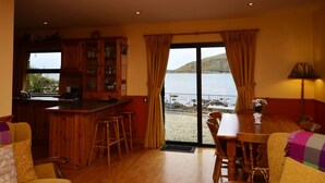 A cozy room with a wooden dining table, chairs, a sofa, and a kitchen area with a view of the sea. - Cottage 108 - Cleggan (Cleggan)