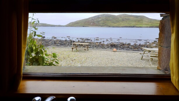 A view from a window overlooking a rocky beach with a wooden bench and a metal fence. - Cottage 108 - Cleggan (Cleggan)
