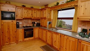 A kitchen with wooden cabinets, a microwave, stove, sink, and a window with curtains. - Cottage 108 - Cleggan (Cleggan)