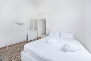 A hotel room with a bed, a nightstand, a wall-mounted towel rack, and a door leading to another room. - UR-NEST Battisti 2 Deluxe (Bologna)