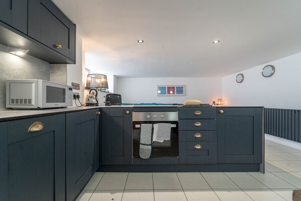 A modern kitchen with dark cabinetry, a microwave, an oven, and a countertop with a kettle. - Mariner’s Belle, Downalong – Sleeps 6 - Off-Site Parking – Dog Friendly (St Ives)