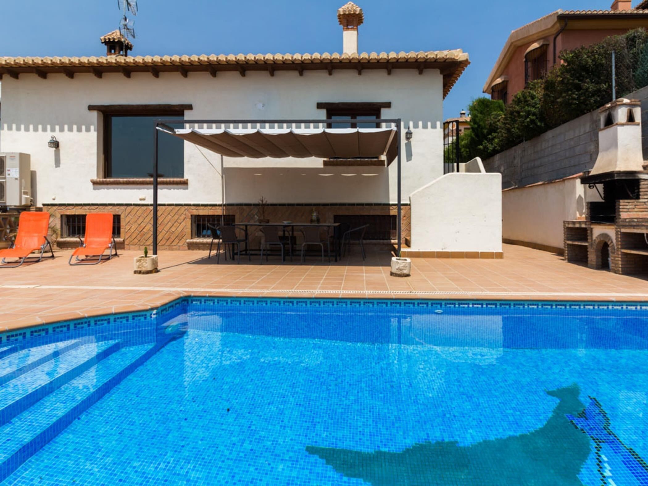 Ein Schwimmbecken mit überdachtem Patio-Bereich, Liegestühlen und einem Grill.