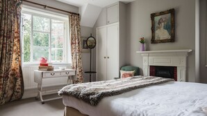 A bedroom with a bed, a dresser, a window with curtains, a fireplace, and a painting on the wall. - A sudeley castle cottage that sleeps 6 guests  in 3 bedrooms (Cheltenham)