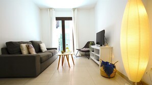 Ein modernes Wohnzimmer mit einer Couch, einem kleinen runden Tisch, einem Fernseher auf einem Schrank und einer Stehlampe.